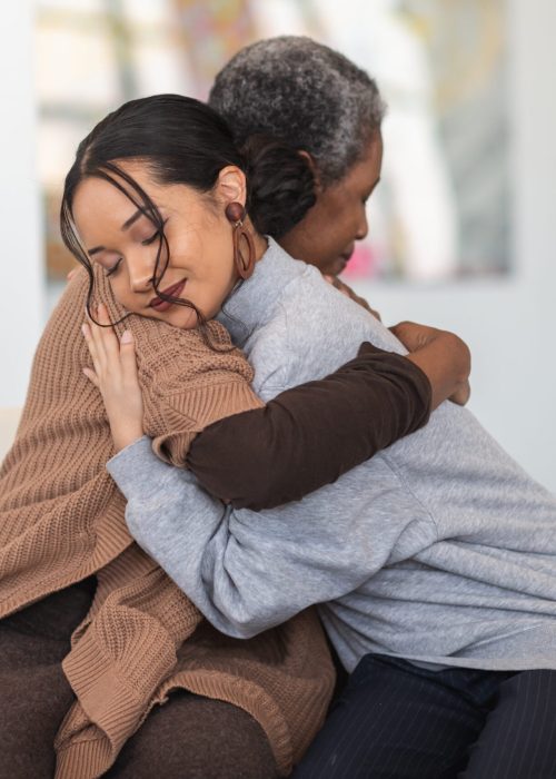 A young mixed-race woman hugs a mature adult black woman. They are sitting next to each other in a medical clinic. The two women are attending a group therapy session. They are showing support and kindness.