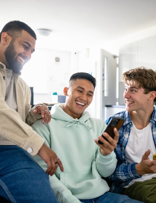 A front view shot of a small group of male uni student friends sitting in their dorm living room, one man is showing his friends something funny on his smartphone and they are laughing together.
